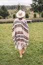Stylish hipster girl in poncho, linen dress and hat walking at lavender field and relaxing in the morning. Bohemian woman enjoying