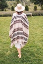 Stylish hipster girl in poncho, linen dress and hat walking at lavender field and relaxing in the morning. Bohemian woman enjoying