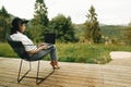 Stylish hipster girl sitting with laptop on terrace with view on woods. Young happy woman in hat using laptop, shopping or working