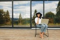 Stylish hipster girl sitting with laptop on wooden porch at big window with trees. Young happy woman in hat using laptop, shopping Royalty Free Stock Photo