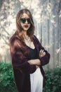 Stylish hipster girl smiling in sunny street on background of wooden wall. Boho girl in cool outfit and sunglasses posing in Royalty Free Stock Photo