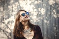 Stylish hipster girl smiling in sunny street on background of wooden wall. Boho girl in cool outfit and sunglasses posing in Royalty Free Stock Photo