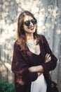 Stylish hipster girl smiling in sunny street on background of wooden wall. Boho girl in cool outfit and sunglasses posing in Royalty Free Stock Photo