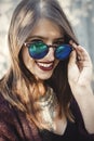 Stylish hipster girl smiling in sunny street on background of wooden wall. Portrait of boho girl in cool outfit and sunglasses Royalty Free Stock Photo
