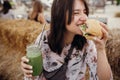 Stylish hipster girl in sunglasses eating delicious vegan burger and holding smoothie in glass jar in hands at street food