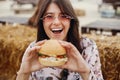 Stylish hipster girl in sunglasses eating delicious vegan burger at street food festival. Happy boho woman tasting and biting