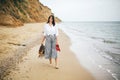 Stylish hipster girl walking barefoot on beach, holding bag and shoes in hand. Happy boho woman relaxing at sea, enjoying walk on Royalty Free Stock Photo