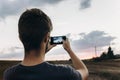 stylish hipster traveler holding smart phone taking photo of beautiful sunset landscape in summer field. instagram photography. e