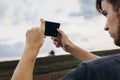 stylish hipster traveler holding smart phone taking photo of beautiful sunset landscape in summer field. instagram photography. e