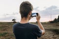 stylish hipster traveler holding smart phone taking photo of beautiful sunset landscape in summer field. instagram photography. e