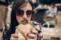 stylish hipster woman eating juicy burger. boho girl biting yummy cheeseburger, smiling at street food festival. summertime. Royalty Free Stock Photo