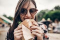 stylish hipster woman eating juicy burger. boho girl biting yummy cheeseburger, smiling at street food festival. summertime. Royalty Free Stock Photo
