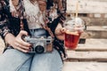 stylish hipster woman holding old photo camera and lemonade. boho girl in denim and bohemian clothes, sitting on wooden bench