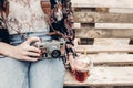 stylish hipster woman holding old photo camera and lemonade. boho girl in denim and bohemian clothes, sitting on wooden bench