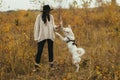 Stylish hipster woman training cute white dog on background of autumn trees. Traveling with pet