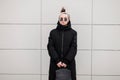 Stylish hipster young woman in stylish sunglasses in a bandana in a fashionable long jacket with a leather stylish backpack near a Royalty Free Stock Photo