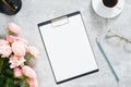 Stylish home office desk with paper clipboard mockup, rose flowers, coffee cup, stationery, glasses on concrete stone background.