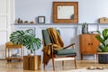 Stylish interior of living room with design armchair, vintage commode, mirror, shelf, tropical leaves, coffee table, plants.