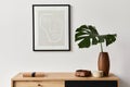 Stylish interior of living room with mock up poster frame, wooden commode, book, tropical leaf in ceramic vase.