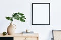 Stylish interior of living room with mock up poster frame, wooden commode, book, tropical leaf in ceramic vase and elegant.