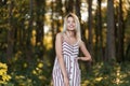 Stylish joyful young woman with a beautiful smile in a trendy pink striped dress standing in the forest on a sunny summer day.