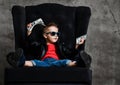 Kid boy in leather jacket is sitting in armchair with a bundle of cash looking at copy space, taking giving a banknote