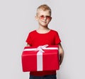 Stylish kid boy in red t-shirt and sunglasses stands holding big present for birthday gift box with ribbon in hands Royalty Free Stock Photo