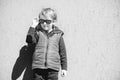 Stylish kid in trendy sunglasses. Kids fashion. Cute little blondy boy in red jacket standing over grey wall outdoors in sunny day