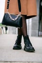 Stylish look. Close-up of a black handbag in the hands. Boots are black. Fashionable girl on the street.Black small handbag. Beige Royalty Free Stock Photo