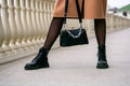 Stylish look. Close-up of a black handbag in the hands. Boots are black. Fashionable girl on the street.Black small handbag. Beige Royalty Free Stock Photo