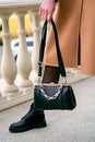 Stylish look. Close-up of a black handbag in the hands. Boots are black. Fashionable girl on the street.Black small handbag. Beige Royalty Free Stock Photo