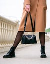 Stylish look. Close-up of a black handbag in the hands. Boots are black. Fashionable girl on the street.Black small handbag. Beige Royalty Free Stock Photo