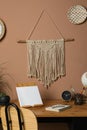 Stylish macrame and wicker wall decor hanging above wooden table with stationery