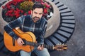 Stylish man with beard in a plaid shirt and jeans in the street near a park with flowers and a guitar in his hands. Macho with a b Royalty Free Stock Photo