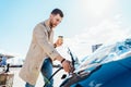 Stylish man with coffe cup in hand inserts plug into the electric car charging socket. Royalty Free Stock Photo