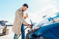Stylish man with coffe cup in hand inserts plug into the electric car charging socket. Royalty Free Stock Photo