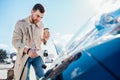 Stylish man with coffe cup in hand inserts plug into the electric car charging socket. Royalty Free Stock Photo
