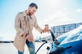 Stylish man with coffe cup in hand inserts plug into the electric car charging socket. Royalty Free Stock Photo