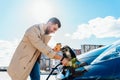 Stylish man with coffe cup in hand inserts plug into the electric car charging socket. Royalty Free Stock Photo
