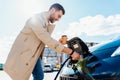 Stylish man with coffe cup in hand inserts plug into the electric car charging socket. Royalty Free Stock Photo