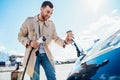 Stylish man with coffe cup in hand inserts plug into the electric car charging socket. Royalty Free Stock Photo