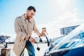 Stylish man with coffe cup in hand inserts plug into the electric car charging socket. Royalty Free Stock Photo