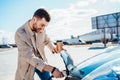 Stylish man with coffe cup in hand inserts plug into the electric car charging socket. Royalty Free Stock Photo