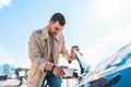 Stylish man with coffe cup in hand inserts plug into the electric car charging socket. Royalty Free Stock Photo