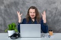 Stylish man with long hair in blue suit striking poses by laptop and accessories
