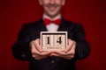 Stylish man in suit hold wooden calendar, set on 14 February with red background, focus on calendar