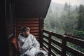 stylish man traveler in blanket relaxing on porch of wooden cabin in rainy day on background of woods in mountains. stylish Royalty Free Stock Photo