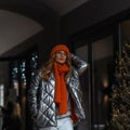 Stylish model of an attractive woman in a winter fashion youth orange-silver outerwear walks around the city near vintage