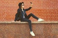 Stylish modern caucasian young woman taking selfie with mobile phone wearing black round hat, jacket and backpack on city street