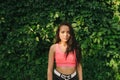 Stylish mulatto girl in a pink T-shirt stands on the background of a green wall covered with a plant, looks into the camera.